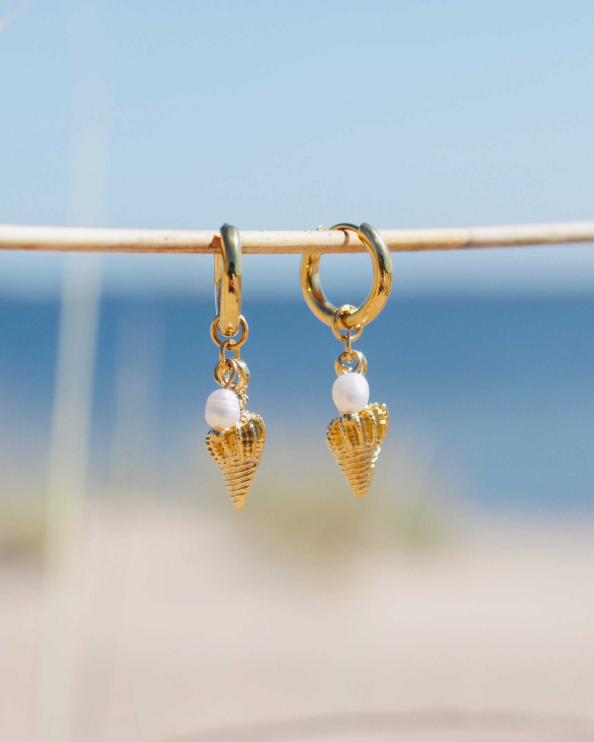 A pair of gold huggie hoop earrings with dangling gold seashell charms and white pearl beads, hanging from a thin wooden stick against a blurred beach background.