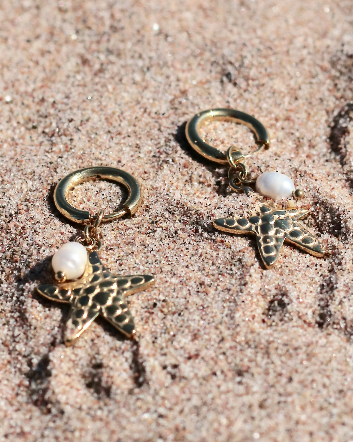 Gold-tone hoop earrings with starfish charms and freshwater pearls displayed on beach sand in Hanko, Finland
