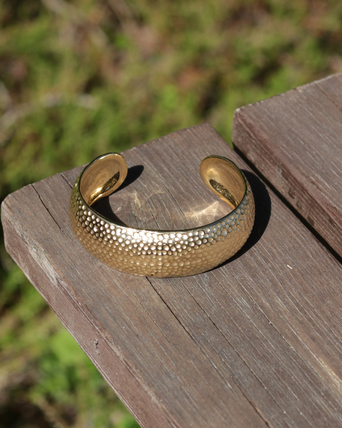 A hammered gold cuff bracelet resting on a weathered wooden board with a blurred green outdoor background.