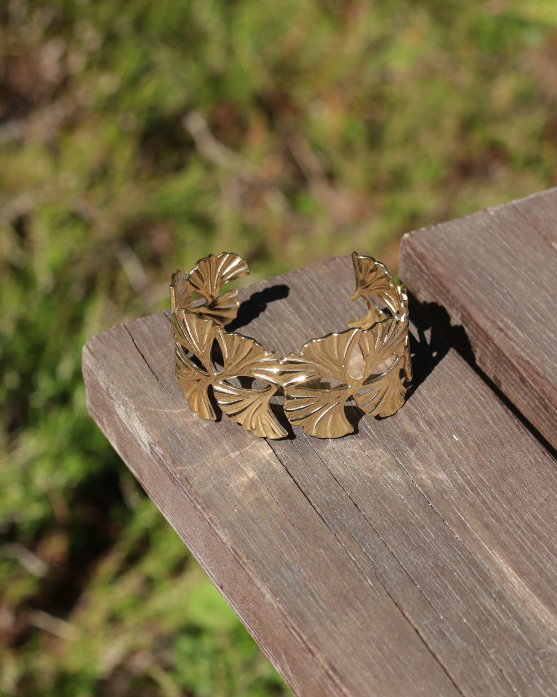A gold-toned metal cuff bracelet with a textured, fan-like leaf pattern, resting on a weathered wooden plank with a blurred green outdoor background.