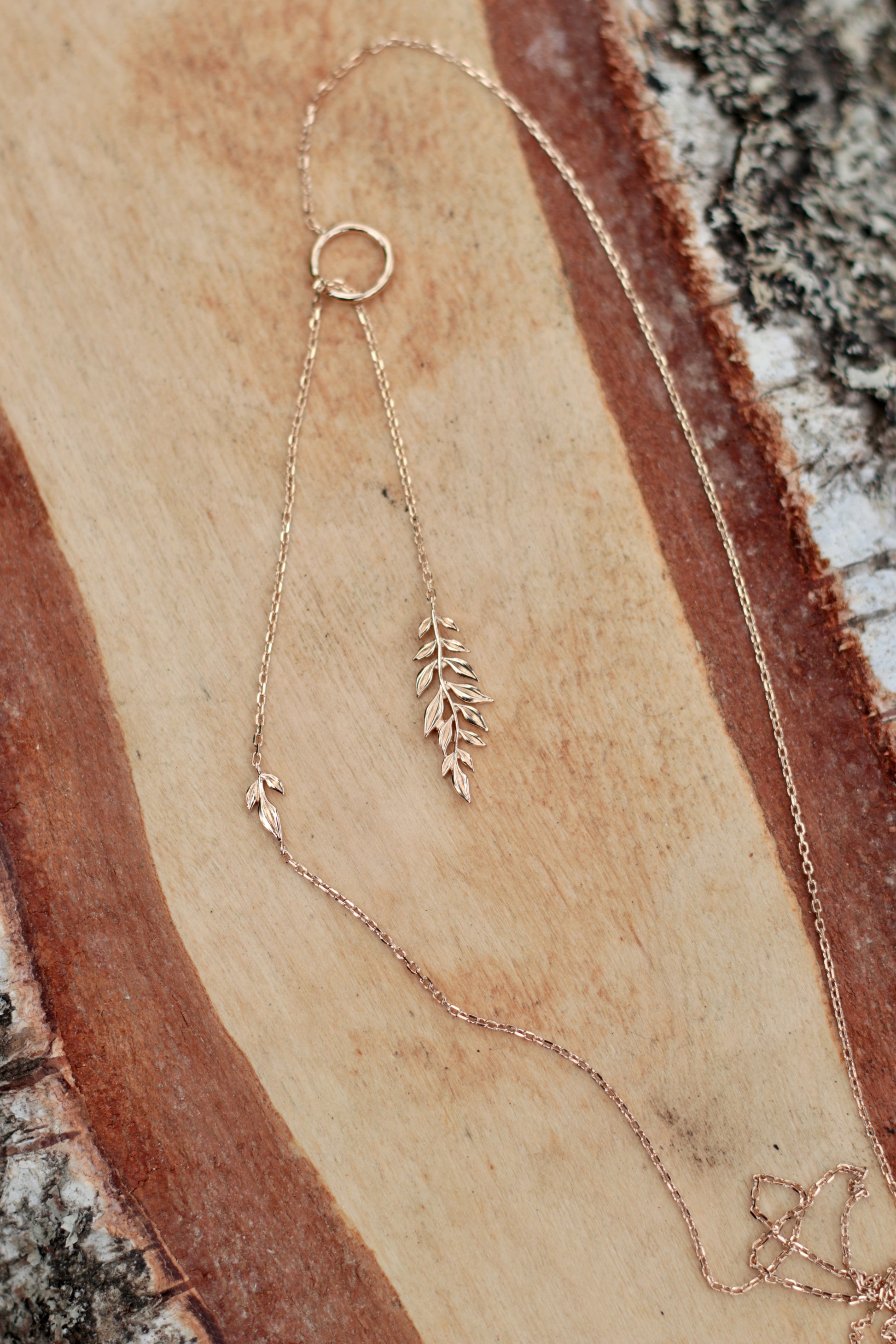 A delicate rose gold lariat necklace with a circular ring, a small leaf charm, and a leafy sprig pendant, displayed on a rustic wood slice background.