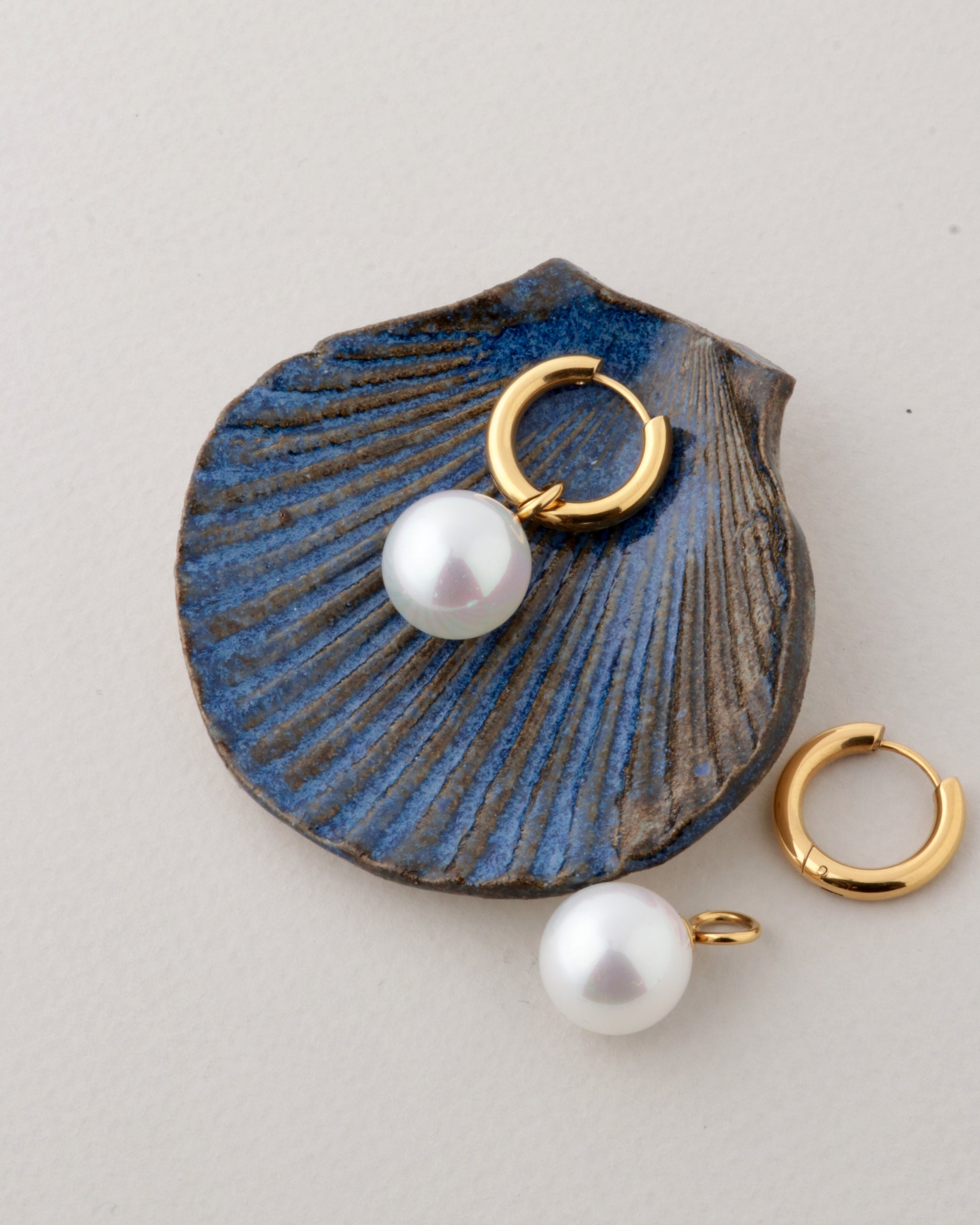 Gold huggie hoop earrings with white freshwater pearls, displayed on a textured blue clam shell prop against a light gray background.