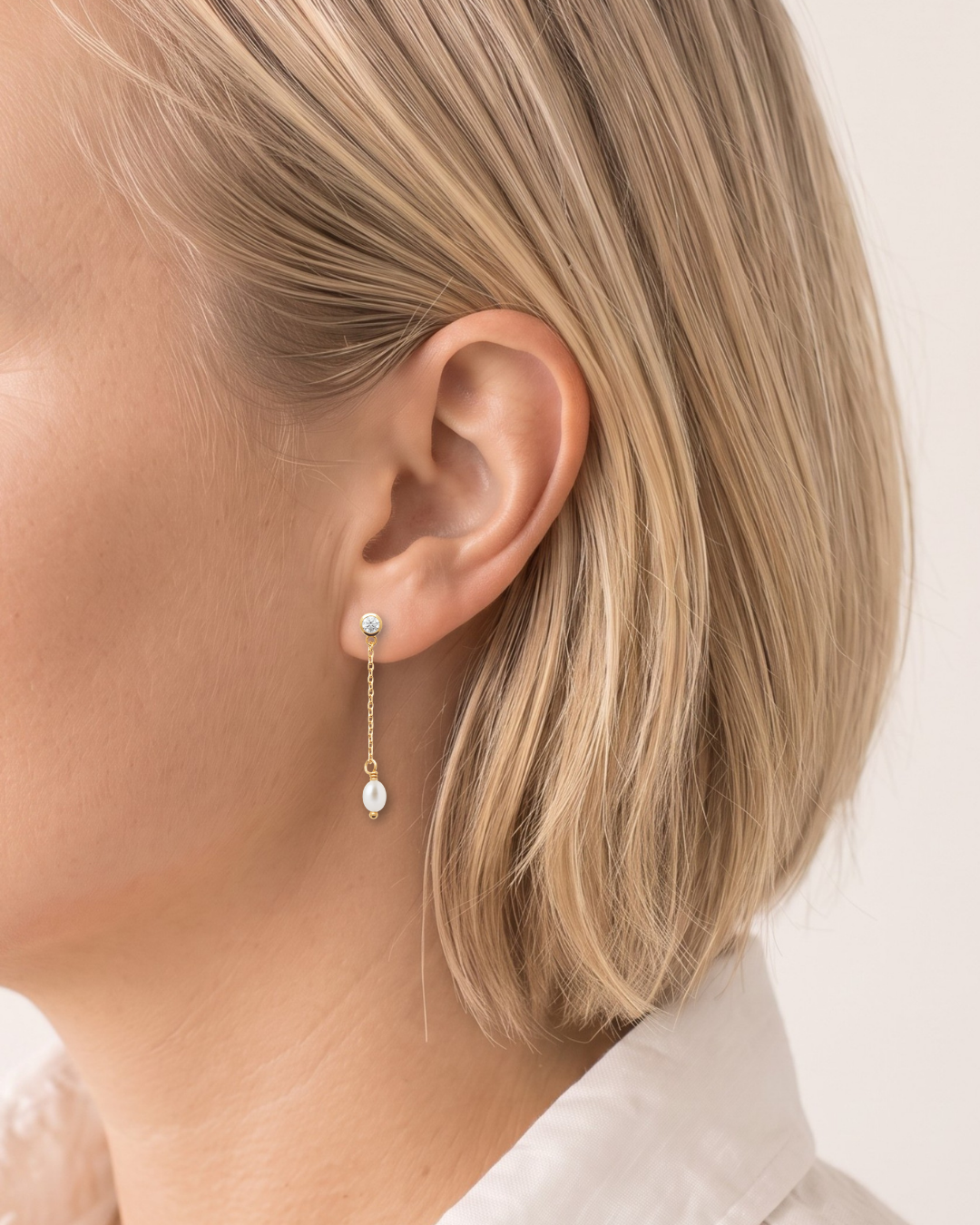 A close-up side profile of a woman with blonde hair wearing a delicate gold dangle earring with a small white pearl stud and a larger teardrop-shaped white pearl drop.