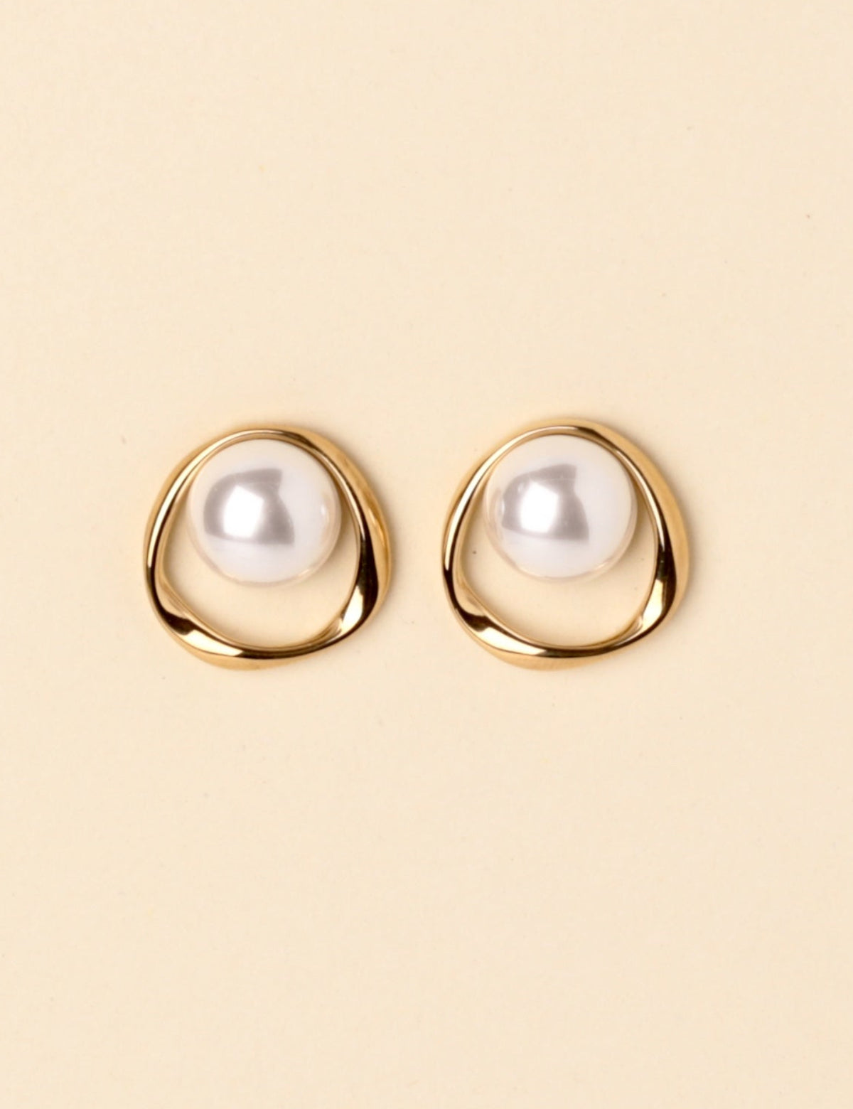 A pair of gold-toned stud earrings with a white pearl set in an organic, open circular shape, displayed on a solid light beige background.