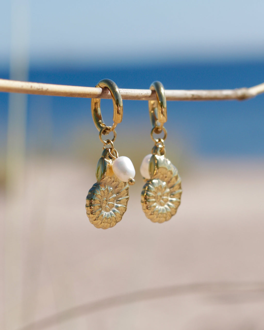 A pair of gold dangle earrings with spiral shell charms and small white pearl beads, hanging from a thin branch with a blurred beach background.