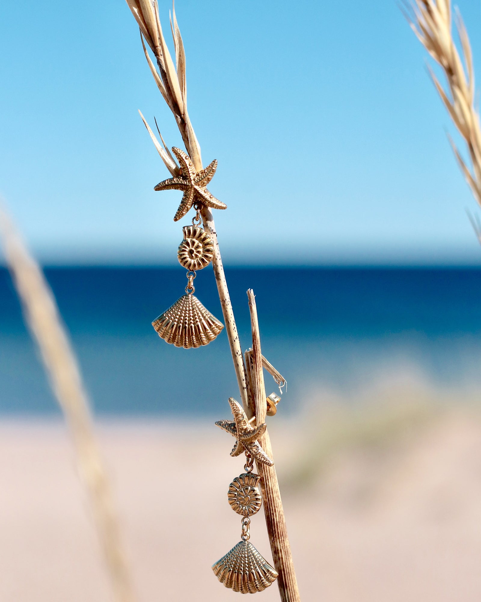 Gold-tone seaside drop earrings with starfish and shell-inspired details photographed by the sea in Hanko, Finland