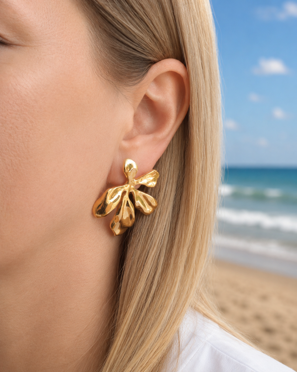 Gold floral earrings worn by a person with a beach in the background