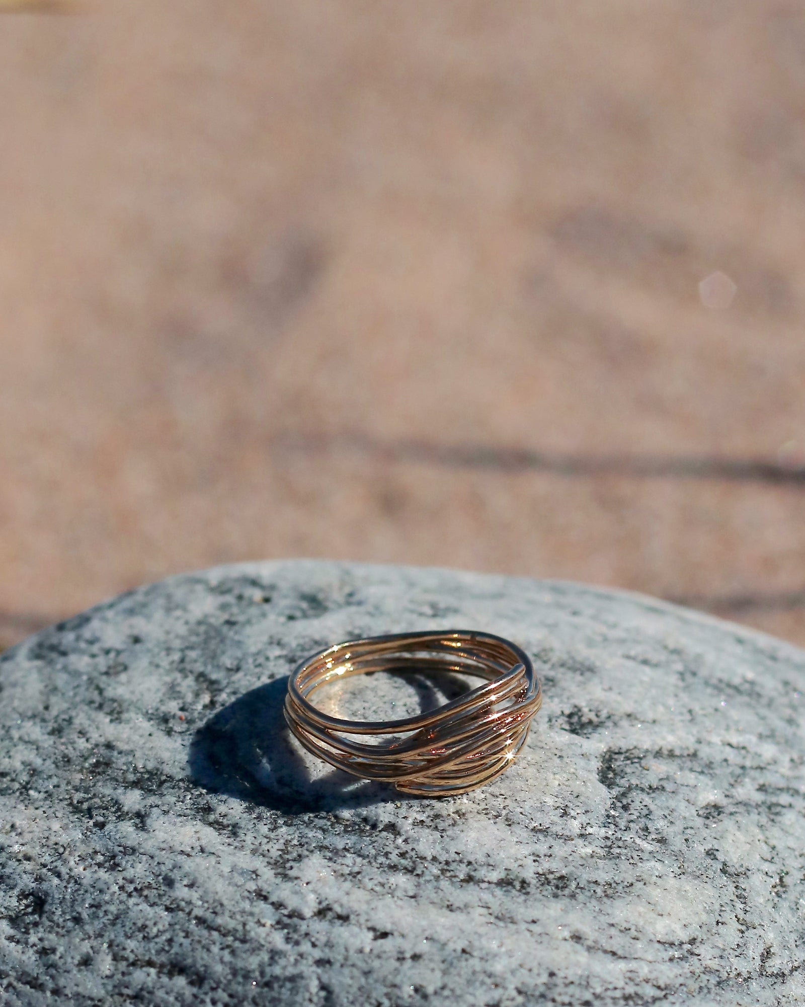 18K gold-plated interwoven band ring with flowing sculptural lines displayed on coastal stones at the beach in Hanko, Finland