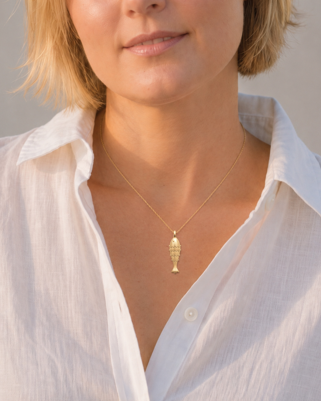 A close-up of a woman wearing a delicate gold chain necklace with a textured fish-shaped pendant, styled with a white linen button-up shirt.