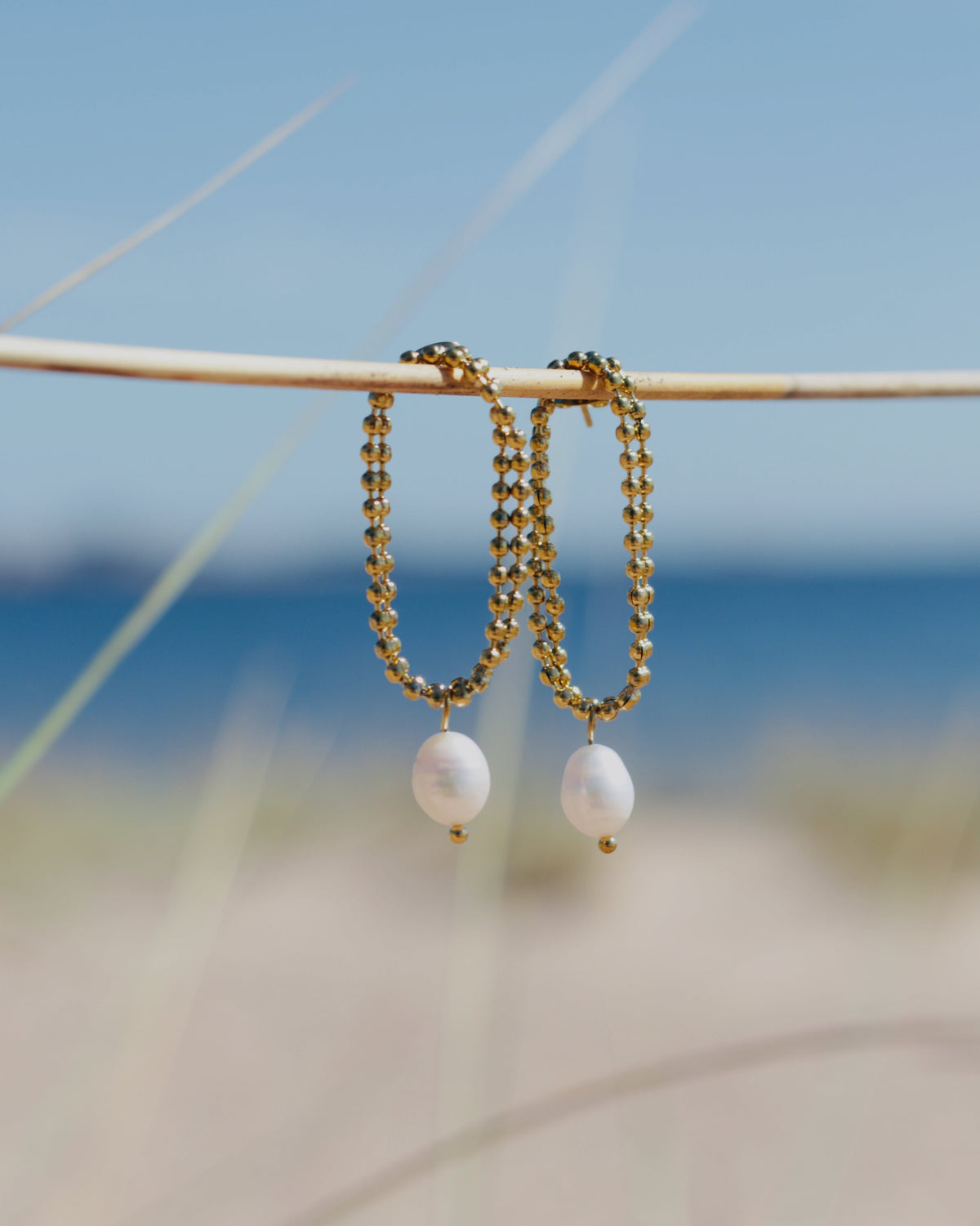 Gold beaded hoop earrings with white pearl dangles hanging from a thin branch, with a blurred beach and blue sky background.
