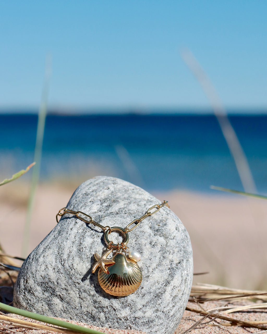 Gold-tone charm necklace with a seashell pendant, starfish charm and freshwater pearl displayed on a beach stone in Hanko, Finland