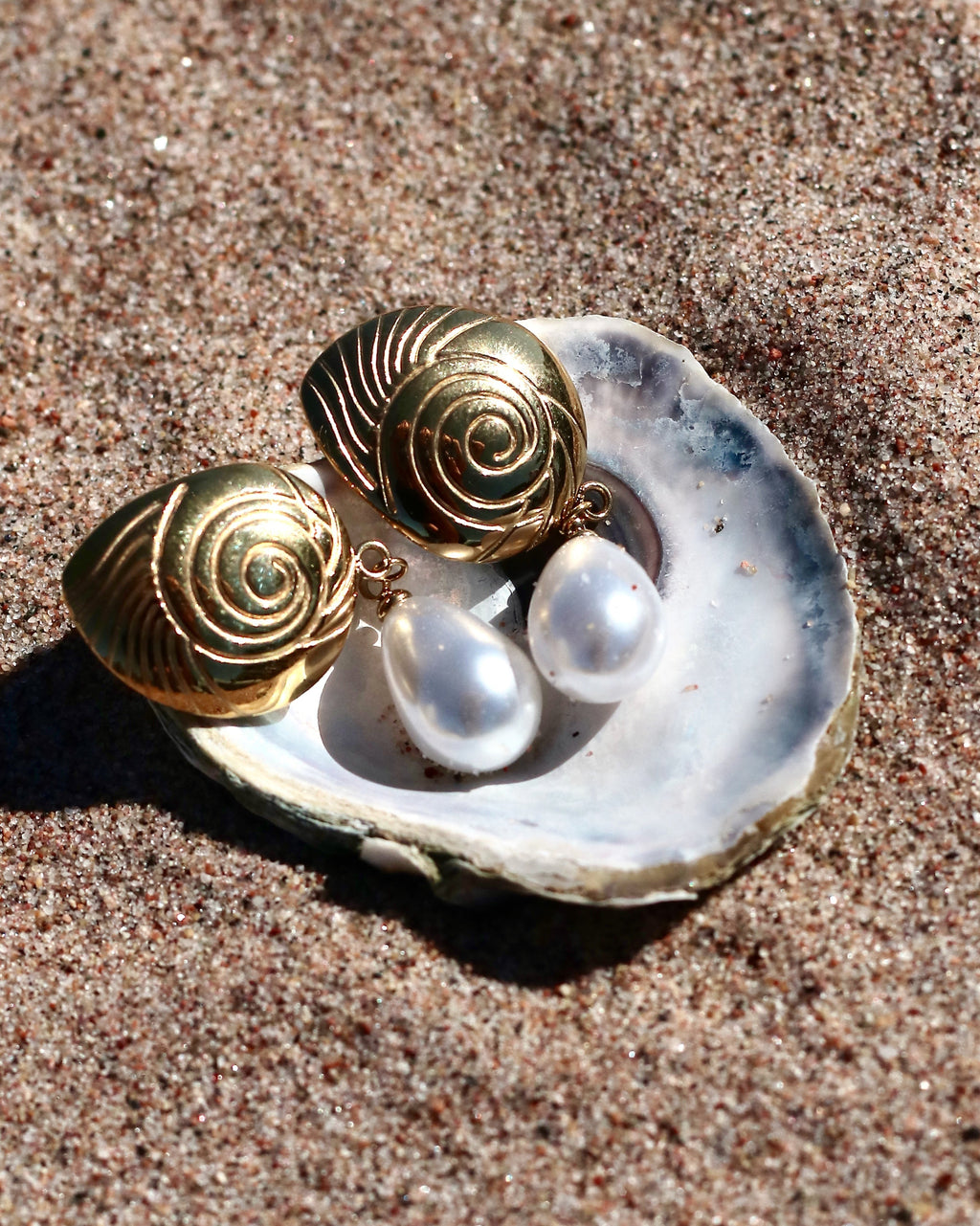 Gold-tone pearl drop earrings with engraved sculptural studs and teardrop pearls displayed in a seashell on the beach in Hanko, Finland